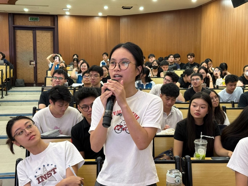 The image depicts a lecture hall with a speaker addressing a large audience, likely a classroom or seminar setting.AI-generated content may be incorrect.